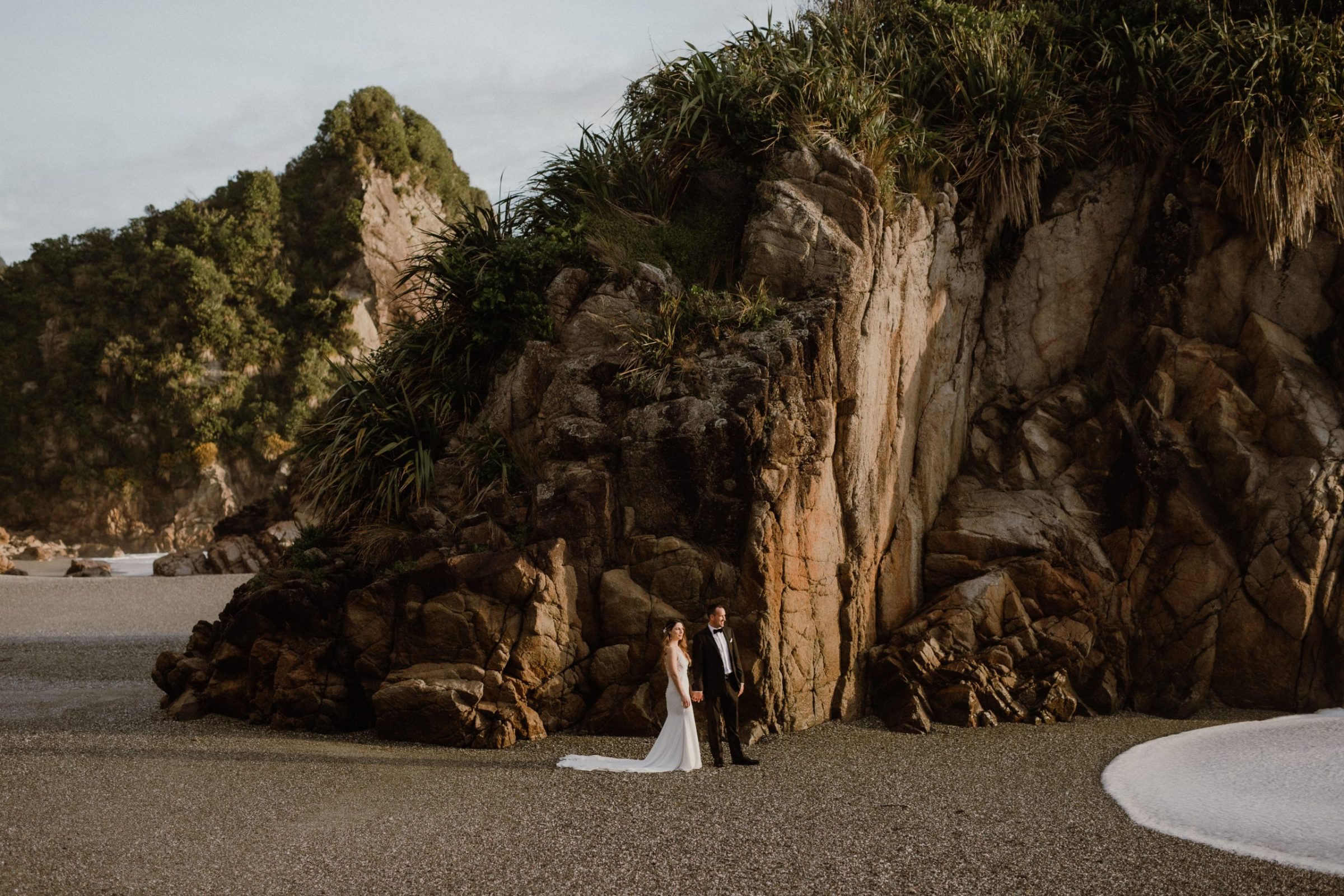 bride and groom during their elopement hold hands on a rocky beach on the West Coast of New Zealand Ana Galloway Photography