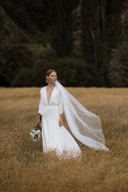 Bride standing in the middle of summery grass field while the breeze passes right through her veil nelson Wedding Ana Galloway Photography