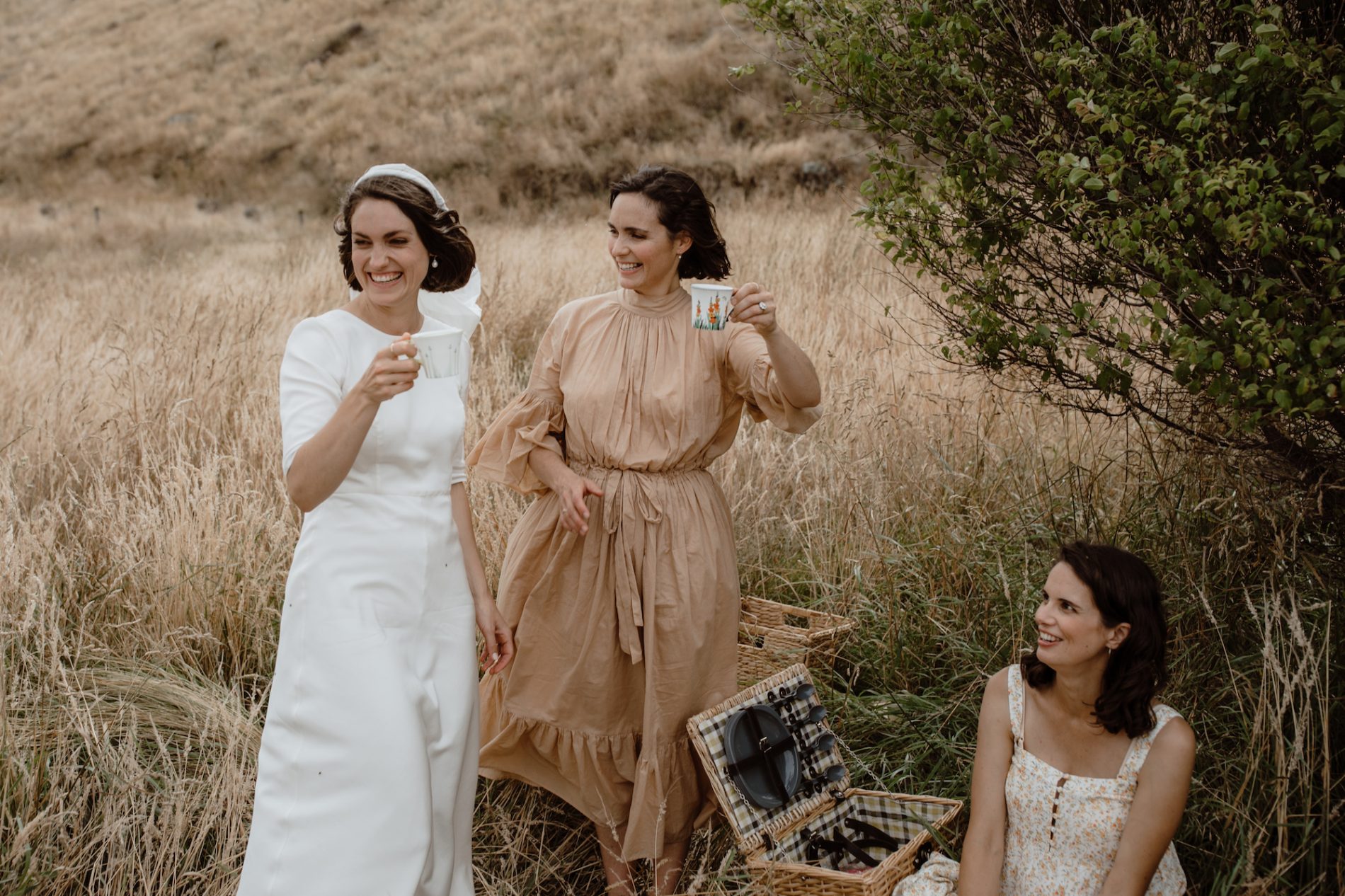 Bride and bridesmaids sharing a picnic with each other in warm tone vintage dresses amongst the soft coastal summer grass Christchurch New Zealand Ana Galloway Photography