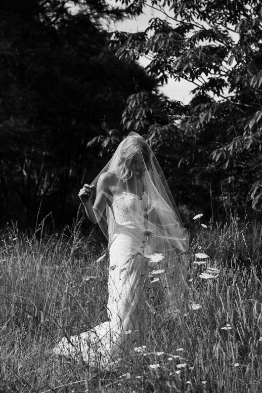 Bride standing amongst long summer grass and wild flowers while she plays with her veil Nelson Wedding Ana Galloway Photography